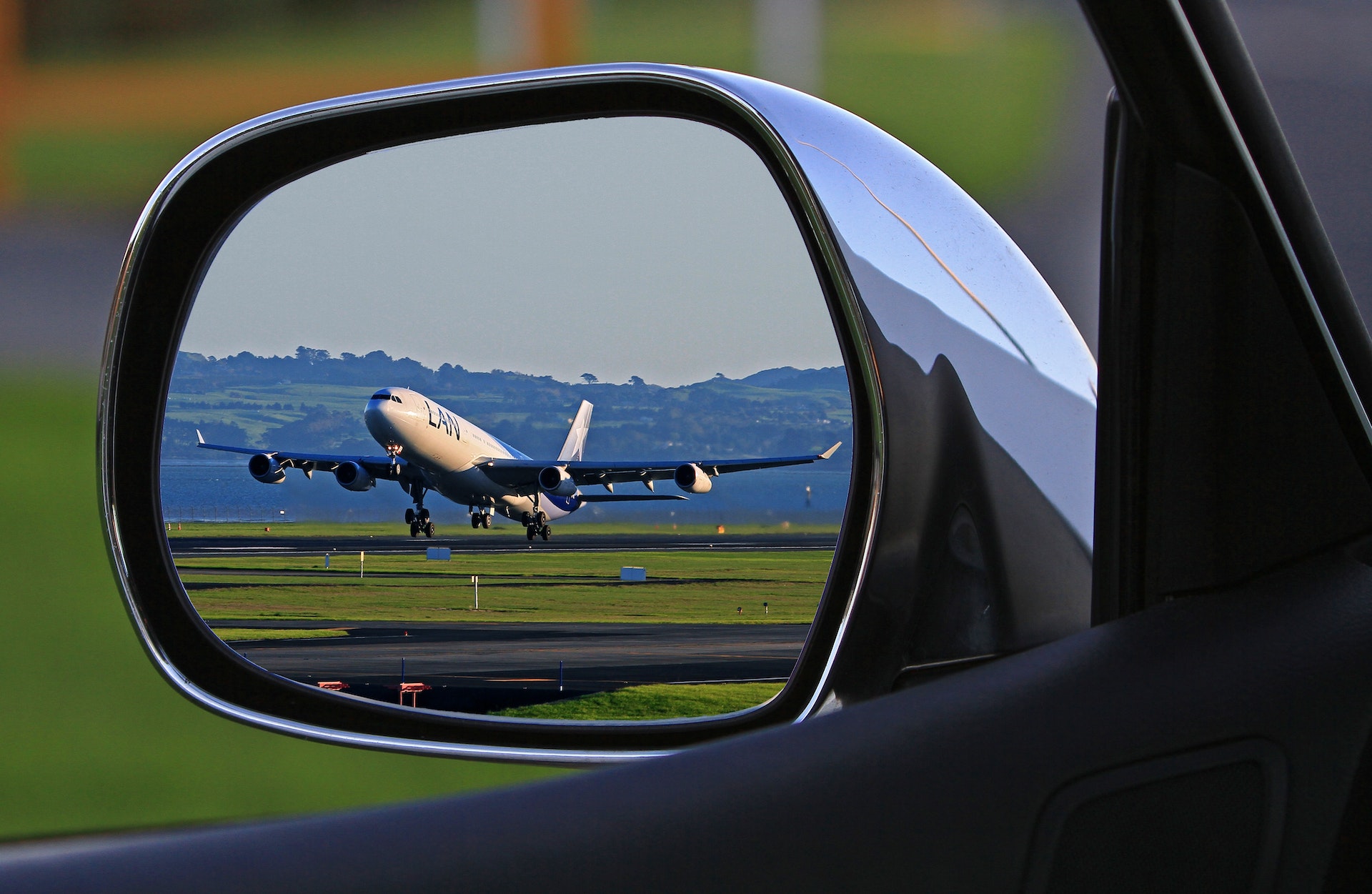 A car's side mirror shows a plane's reflection, linking the skies from Liverpool to Manchester