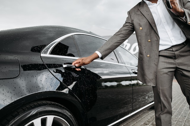 Businessman in a grey suit opening the door of a sleek black car, representing luxury and executive services available for airport taxi booking.